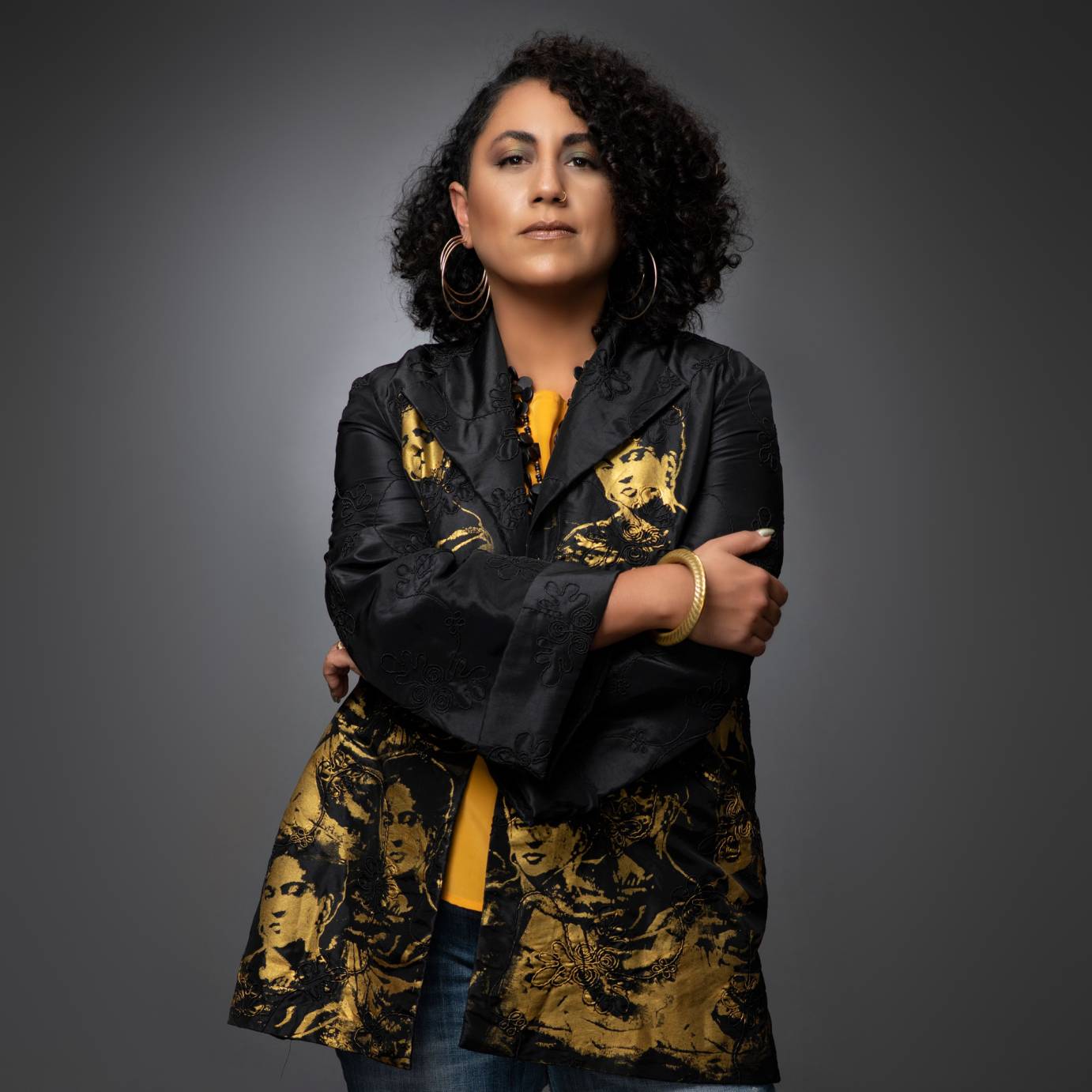 photo of a light brown skinned woman composer Taina Asili staring at us. She wears large hoop earrings a bright yellow shirt and blue jeans topped off by a loose fitting black jacket decorated with gold images of Frida Kahlo. 